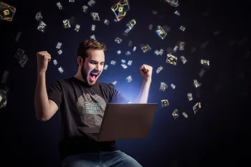 A man celebrating a winning moment on his laptop while gaming symbols float around him, capturing the excitement of OKTA games.